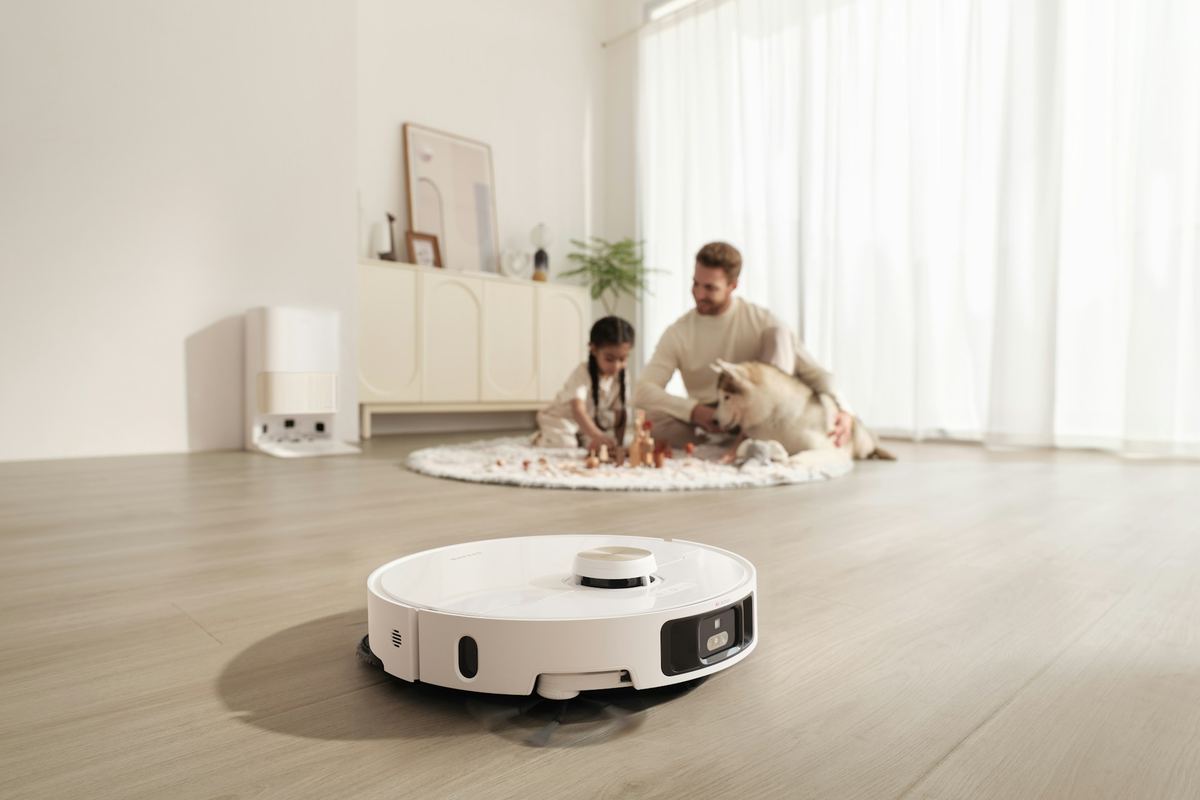 A white robot vacuum on a wooden floor while a parent and child play with a dog on a rug behind it