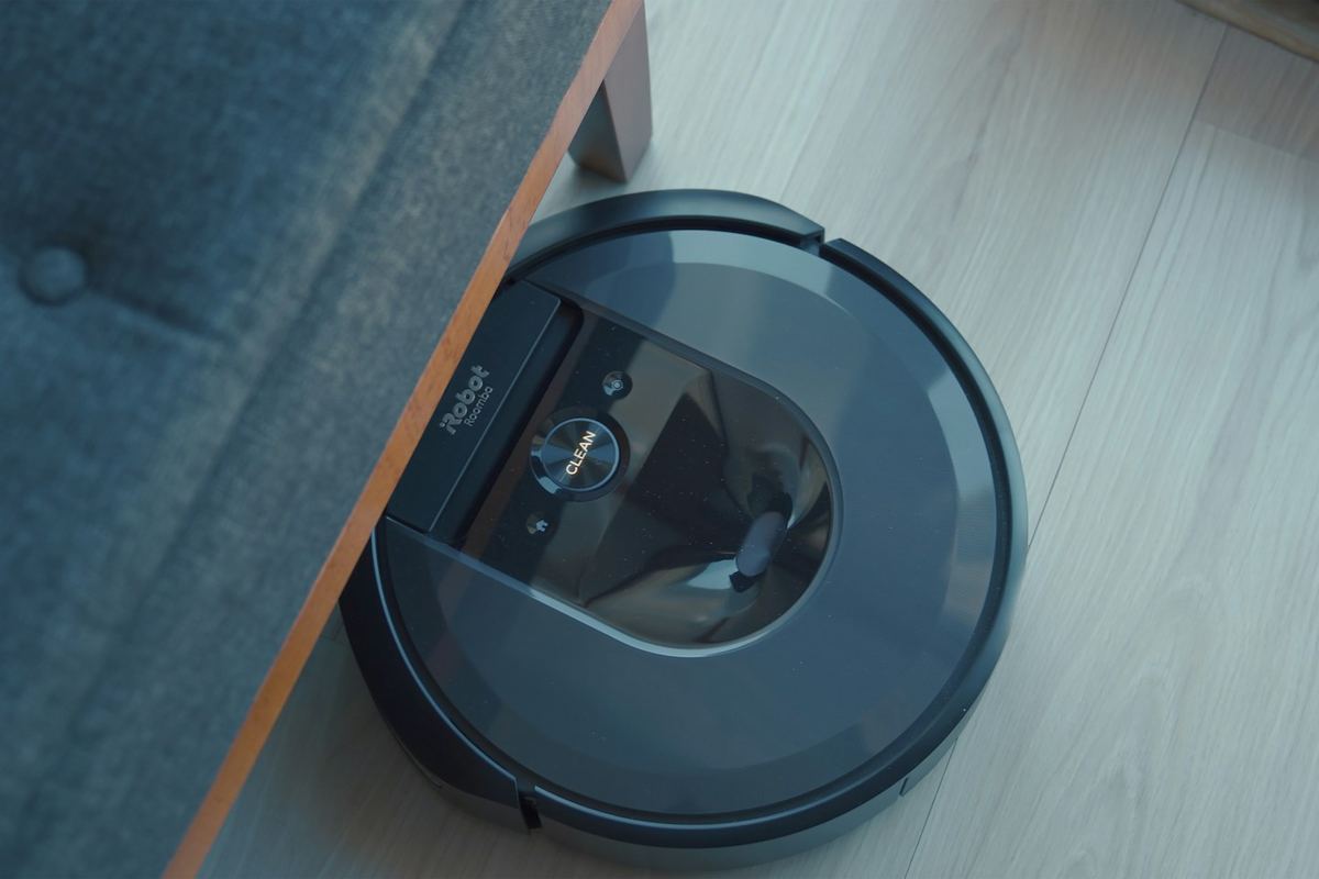 A dark robot vacuum partly under the edge of a blue sofa on a wooden floor
