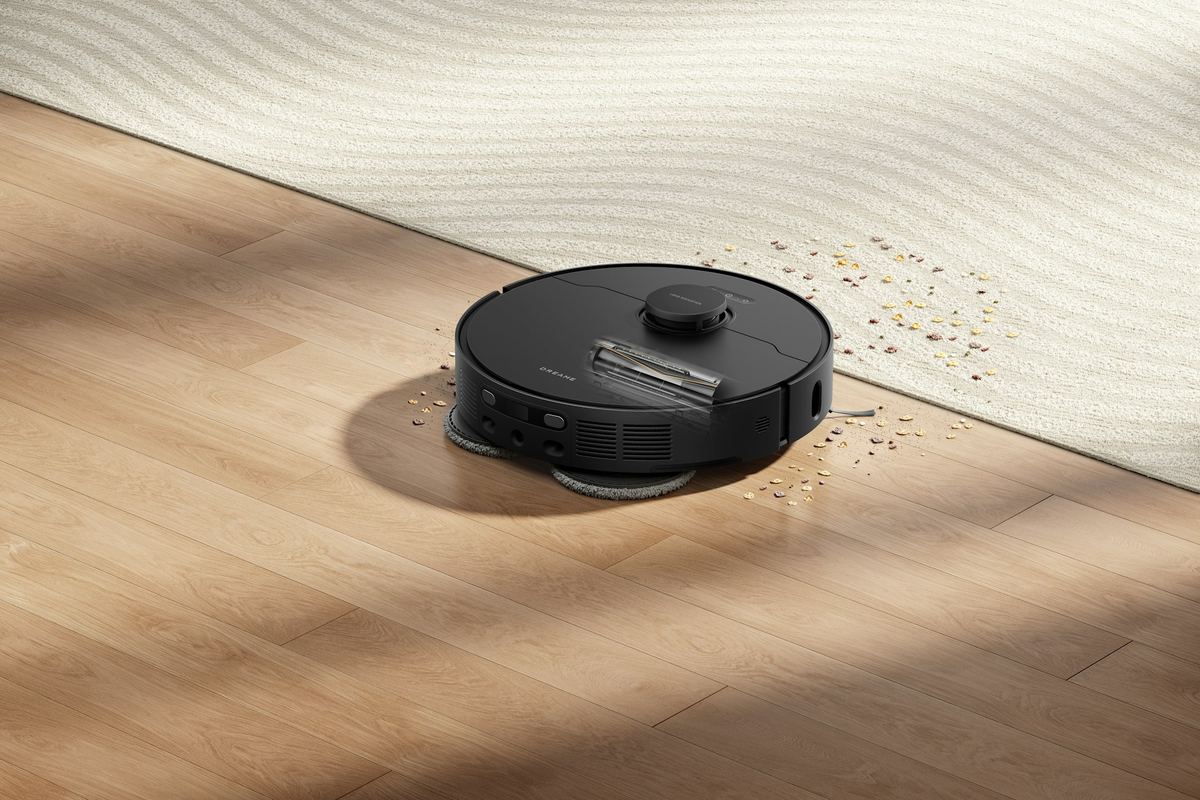 A black robot vacuum on a wooden floor next to scattered crumbs and a cream rug