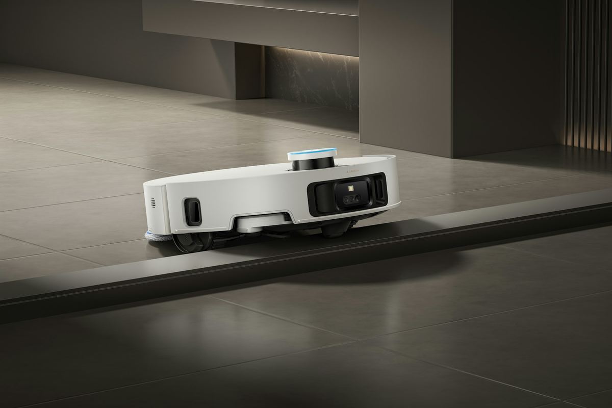 A white robot vacuum with its front wheels raised as it crosses a step from a wooden floor onto tile